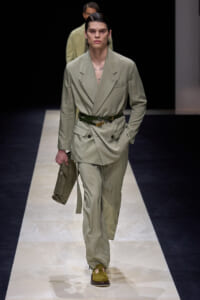 Male model on a runway wearing a sage green double-breasted suit with a dark green belt, holding a matching clutch in one hand.