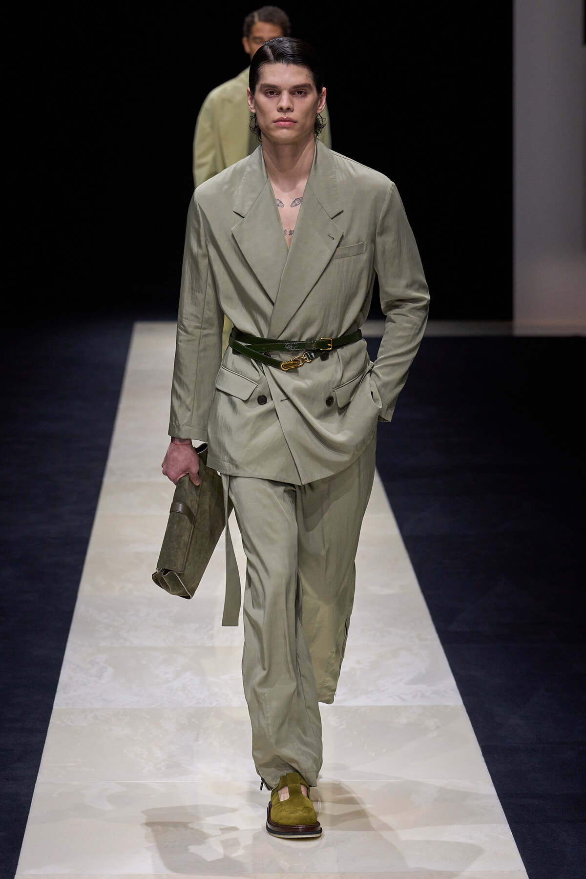 Male model on a runway wearing a sage green double-breasted suit with a dark green belt, holding a matching clutch in one hand.