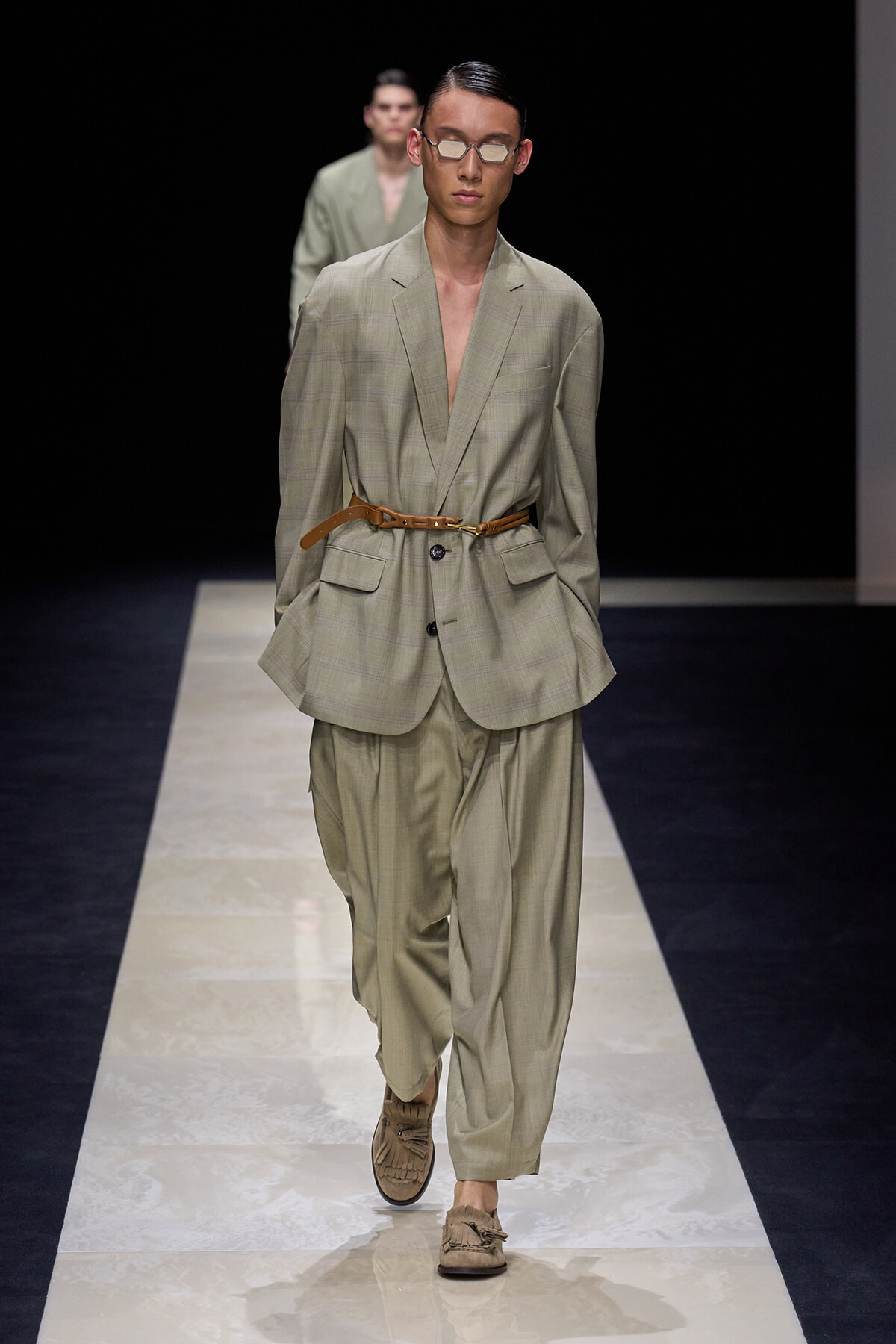Male model on a runway wearing a light beige checked suit with a brown belt and loafers, plus sunglasses, mid-stride.