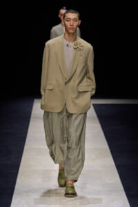 Male model walks a fashion runway in a beige oversized blazer with a flower brooch, taupe top, and light plaid trousers with green loafers.
