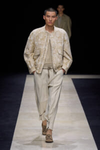 Male model walks the runway in a beige textured jacket over a mesh knit top and pale wide pants, with his hands in pockets.