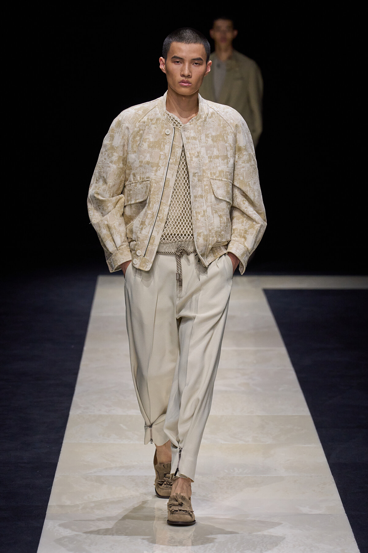 Male model walks the runway in a beige textured jacket over a mesh knit top and pale wide pants, with his hands in pockets.