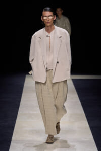 Model on a runway wearing an oversized pale blazer over a light top and wide beige striped trousers, with tan sneakers and tinted sunglasses.