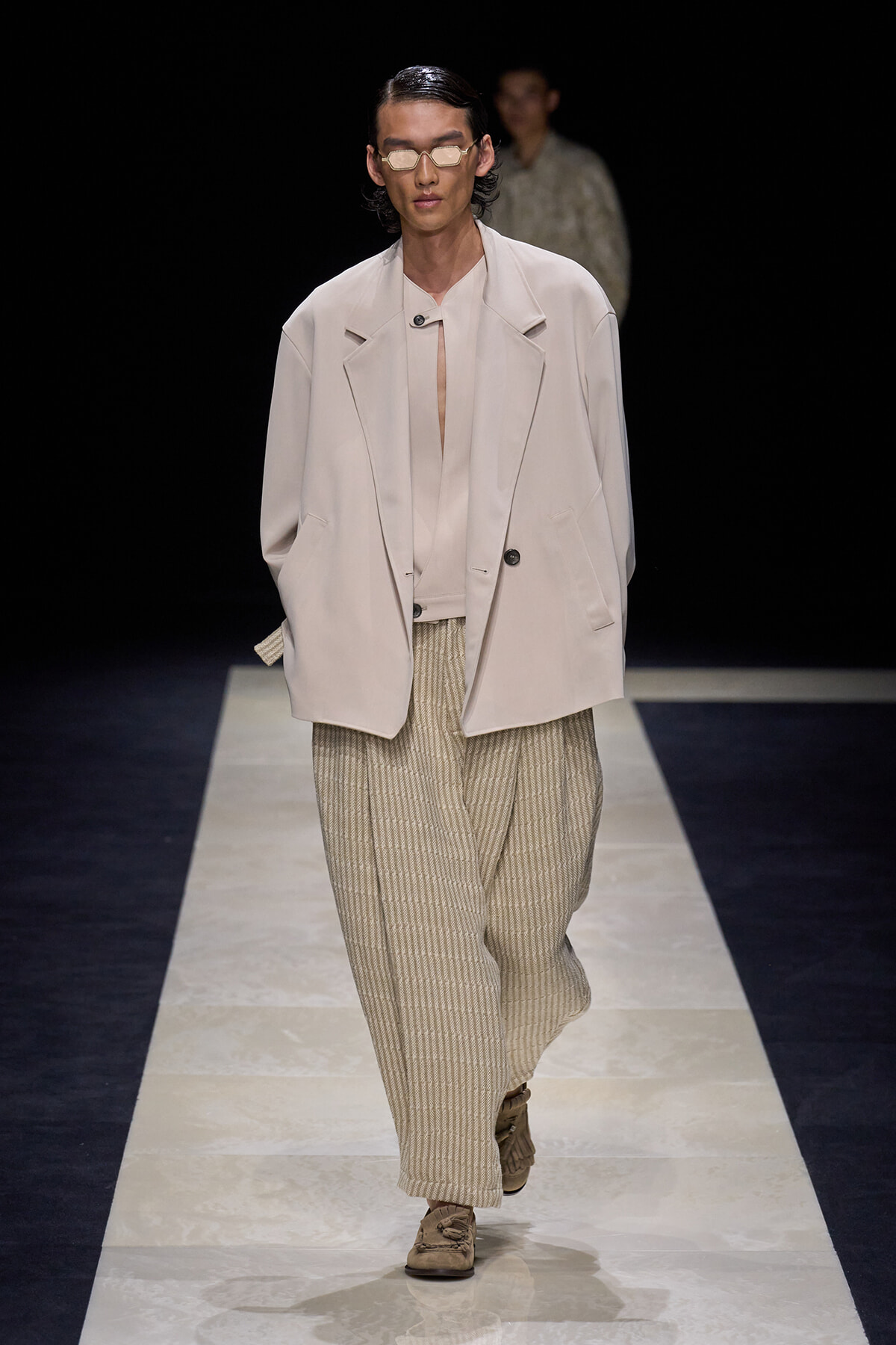 Model on a runway wearing an oversized pale blazer over a light top and wide beige striped trousers, with tan sneakers and tinted sunglasses.