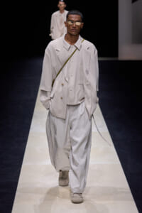 Male model on a runway wearing an oversized beige suit with a crossbody strap and pale sneakers, sunglasses.
