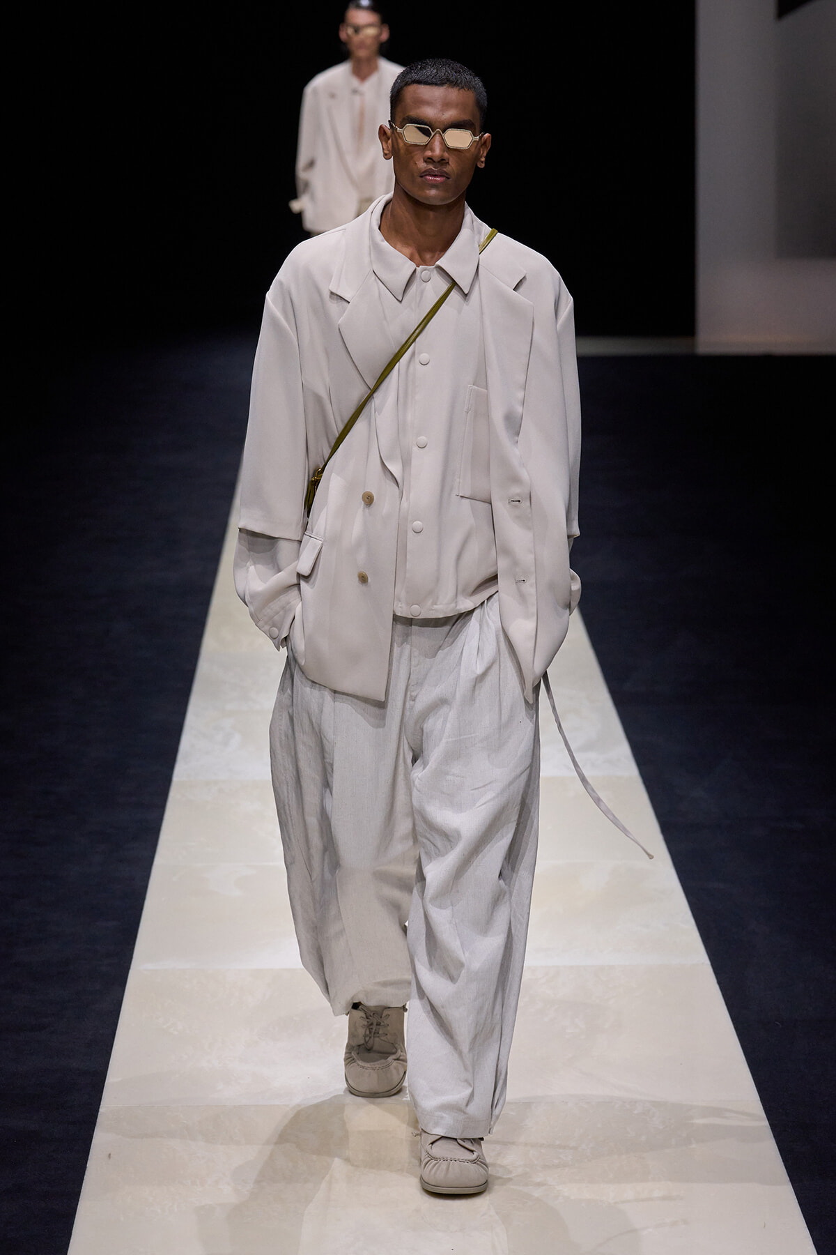 Male model on a runway wearing an oversized beige suit with a crossbody strap and pale sneakers, sunglasses.
