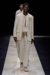 Male model on a runway wearing an oversized off-white blazer with a pale pink shirt, matching wide trousers, floral lapel pin, patterned belt, and light sneakers.