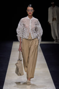 Model walks a runway in a sheer white embroidered blouse and loose beige trousers, carrying a beige tote bag and wearing sunglasses.