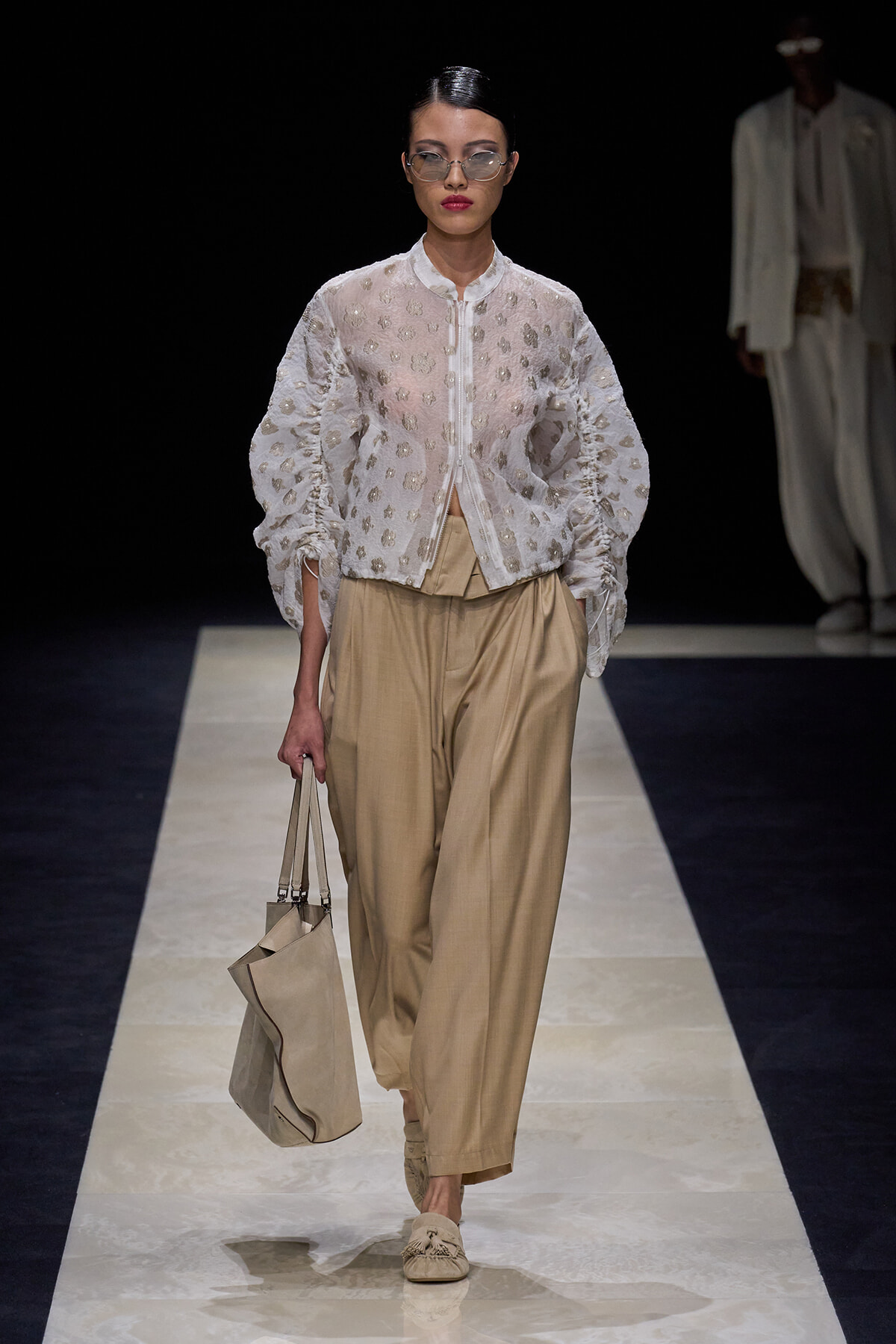 Model walks a runway in a sheer white embroidered blouse and loose beige trousers, carrying a beige tote bag and wearing sunglasses.