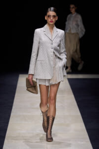 Model walks a fashion runway in a light patterned blazer with matching shorts, beige clutch, and lace-up knee-high boots.