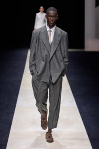 Male model walks a gray oversized double-breasted suit with a white shirt and beige tie on a fashion runway, hands in pockets, brown loafers visible.
