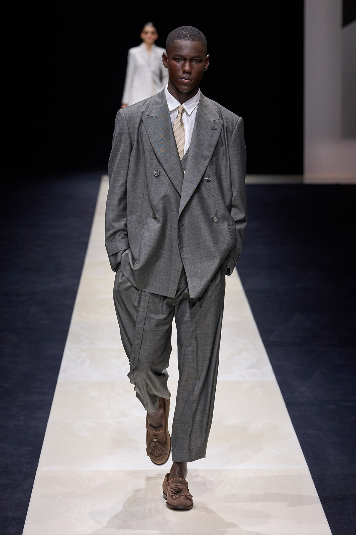 Male model walks a gray oversized double-breasted suit with a white shirt and beige tie on a fashion runway, hands in pockets, brown loafers visible.