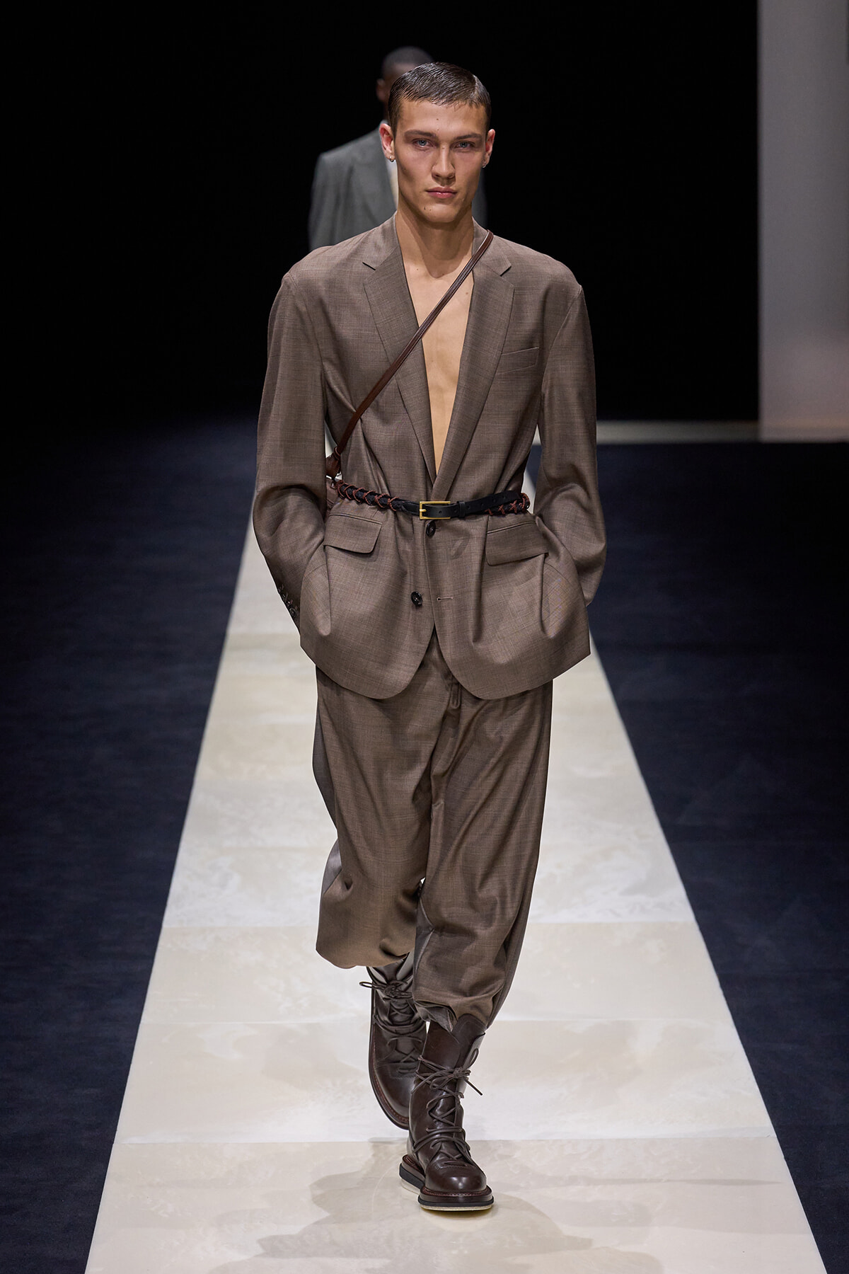 Male model walks a fashion runway in a taupe suit with an oversized blazer, open chest, braided belt, and brown lace-up boots.