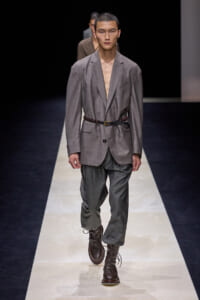 Male model walking a runway in a gray suit with a braided belt and brown lace-up boots.
