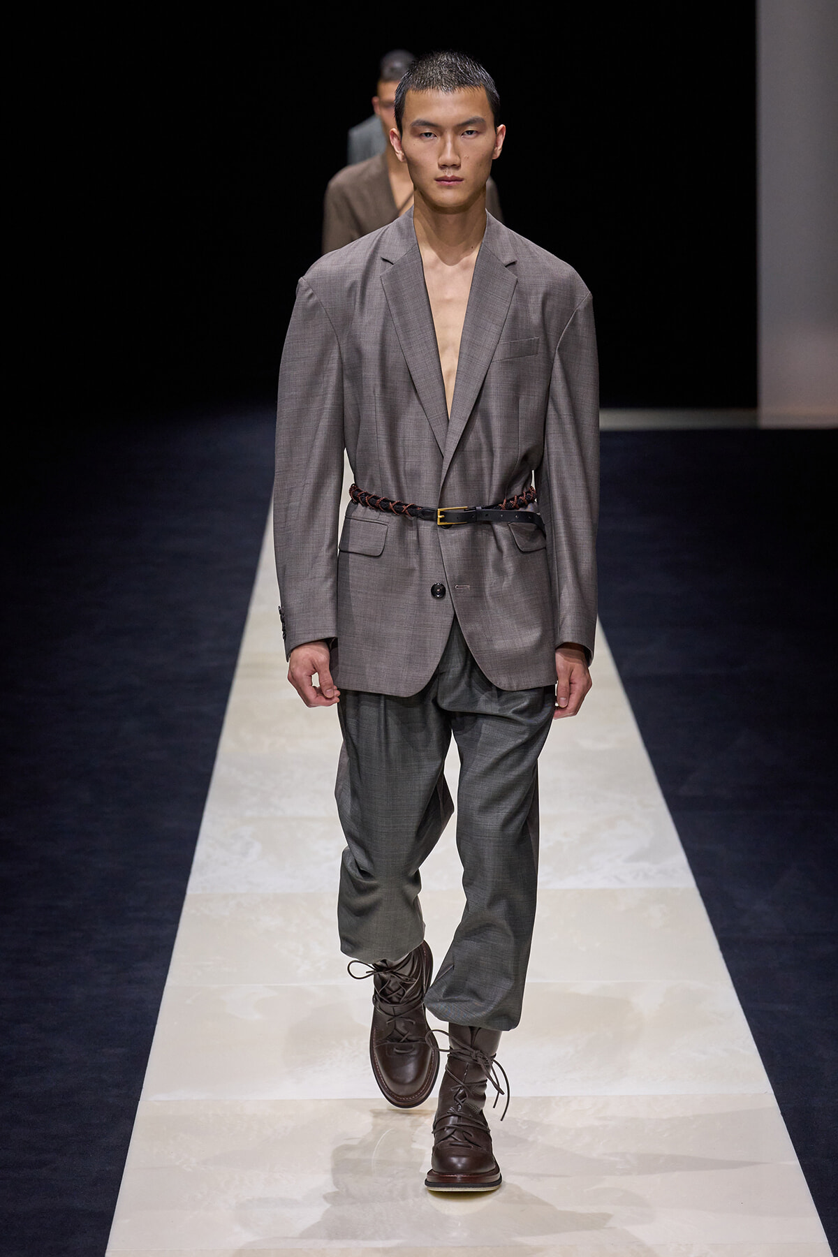 Male model walking a runway in a gray suit with a braided belt and brown lace-up boots.