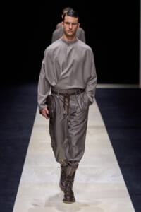 Male fashion model on a runway wearing a gray loose-fit tunic and matching pants with a braided belt, brown lace-up boots, and a brown clutch in hand.