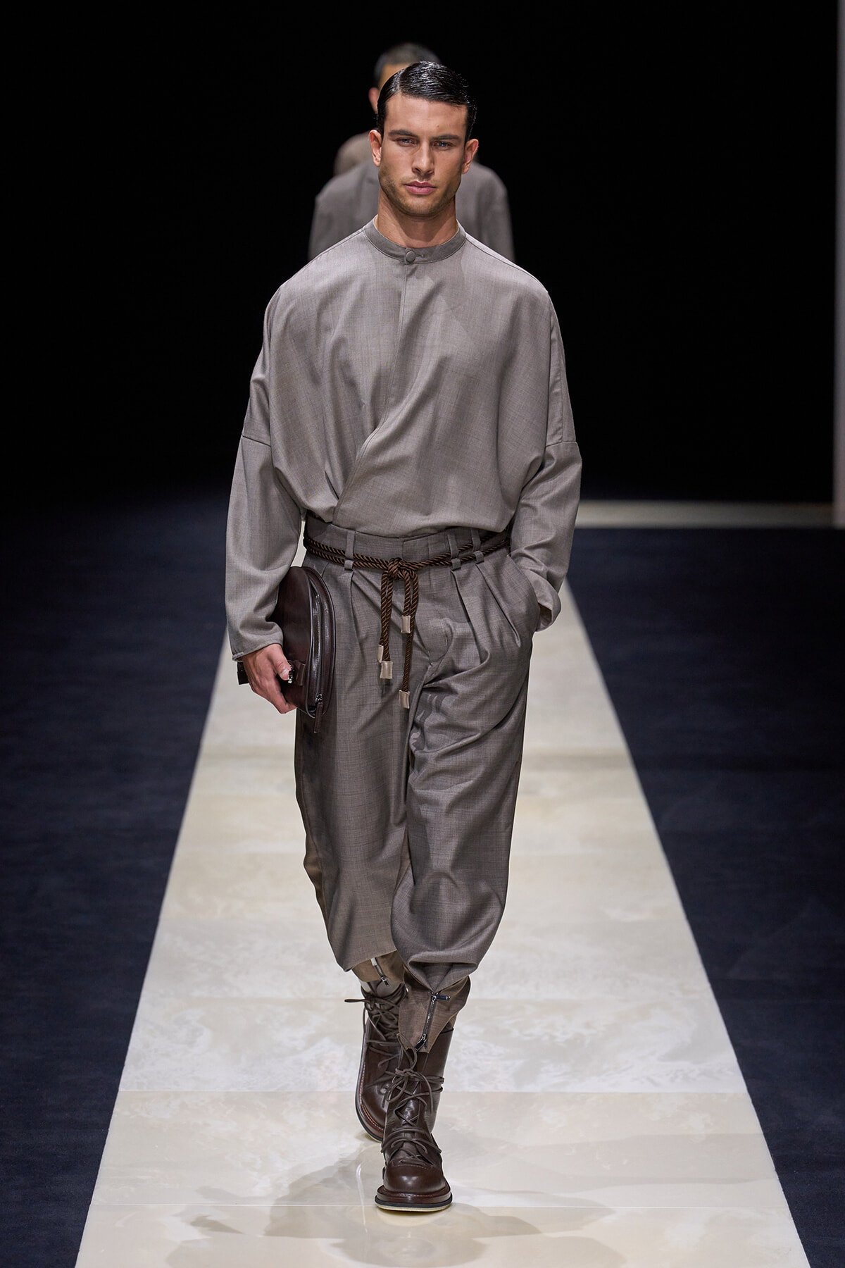 Male fashion model on a runway wearing a gray loose-fit tunic and matching pants with a braided belt, brown lace-up boots, and a brown clutch in hand.
