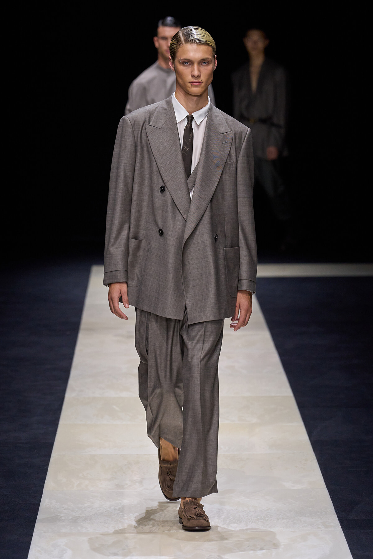 Male model walks a gray oversized double-breasted suit on a runway, white shirt and dark tie, brown loafers, dark backdrop behind.