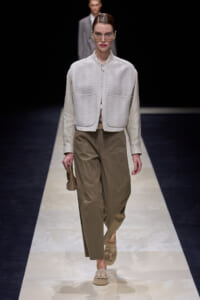 Model walking the runway in a light gray quilted zip-front cropped vest over a beige shirt, and taupe wide-leg trousers with beige loafers, holding a small beige clutch.