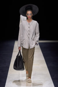 Model walking a fashion runway wearing a light gray, button-front jacket with large pockets, olive trousers, oversized black hat, sunglasses, and carrying a black woven tote.