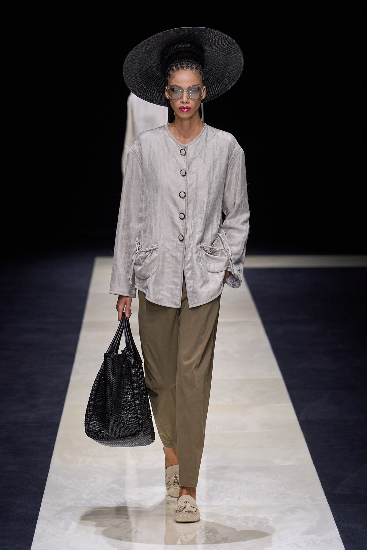 Model walking a fashion runway wearing a light gray, button-front jacket with large pockets, olive trousers, oversized black hat, sunglasses, and carrying a black woven tote.