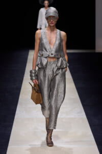 Model on a runway wearing a silver textured sleeveless vest with matching pants, cap, oversized sunglasses, and a small brown handbag.