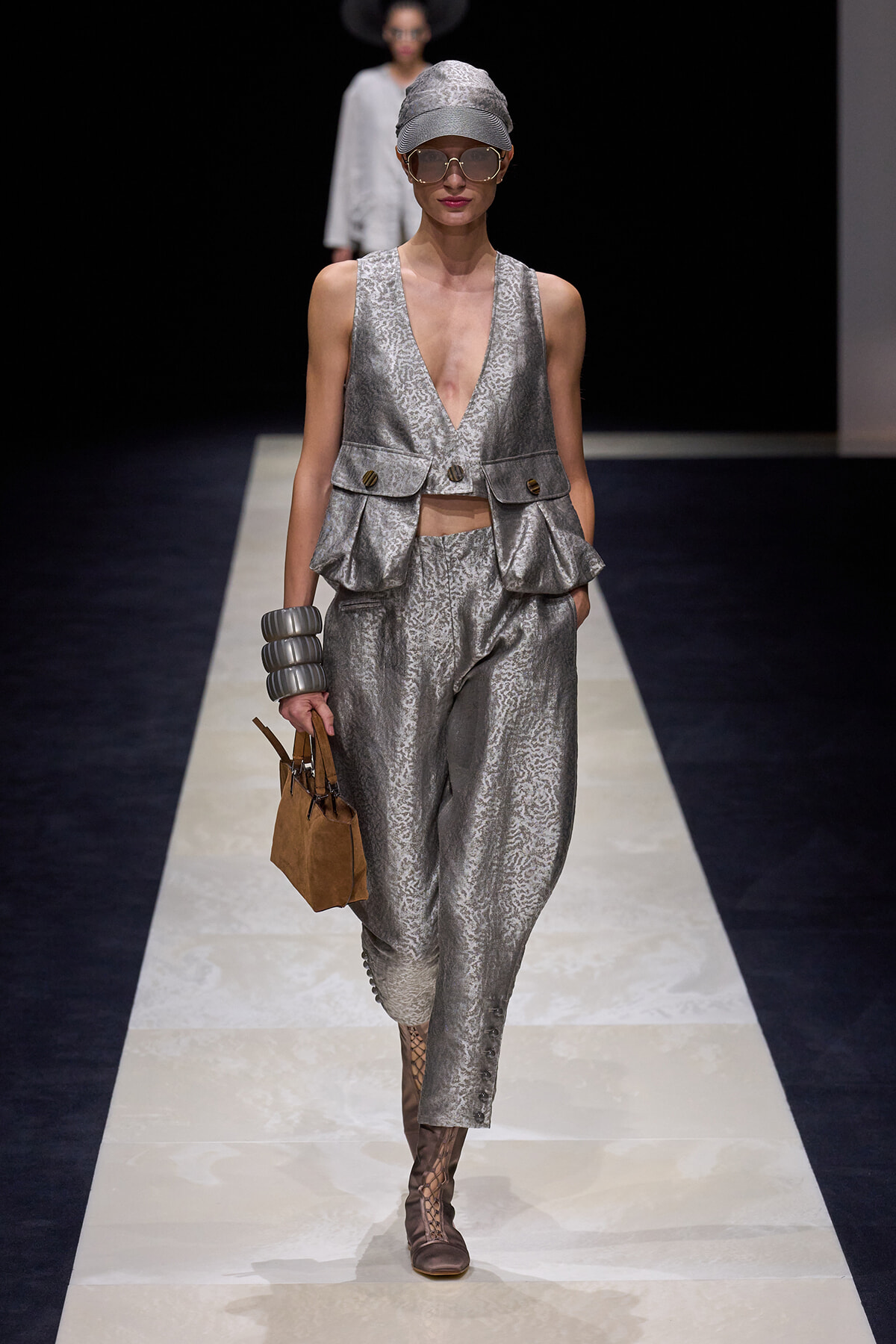 Model on a runway wearing a silver textured sleeveless vest with matching pants, cap, oversized sunglasses, and a small brown handbag.