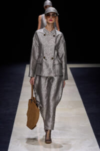 Model on a runway wearing a metallic silver textured suit with cap, sunglasses, and a brown handbag of tan suede kat.