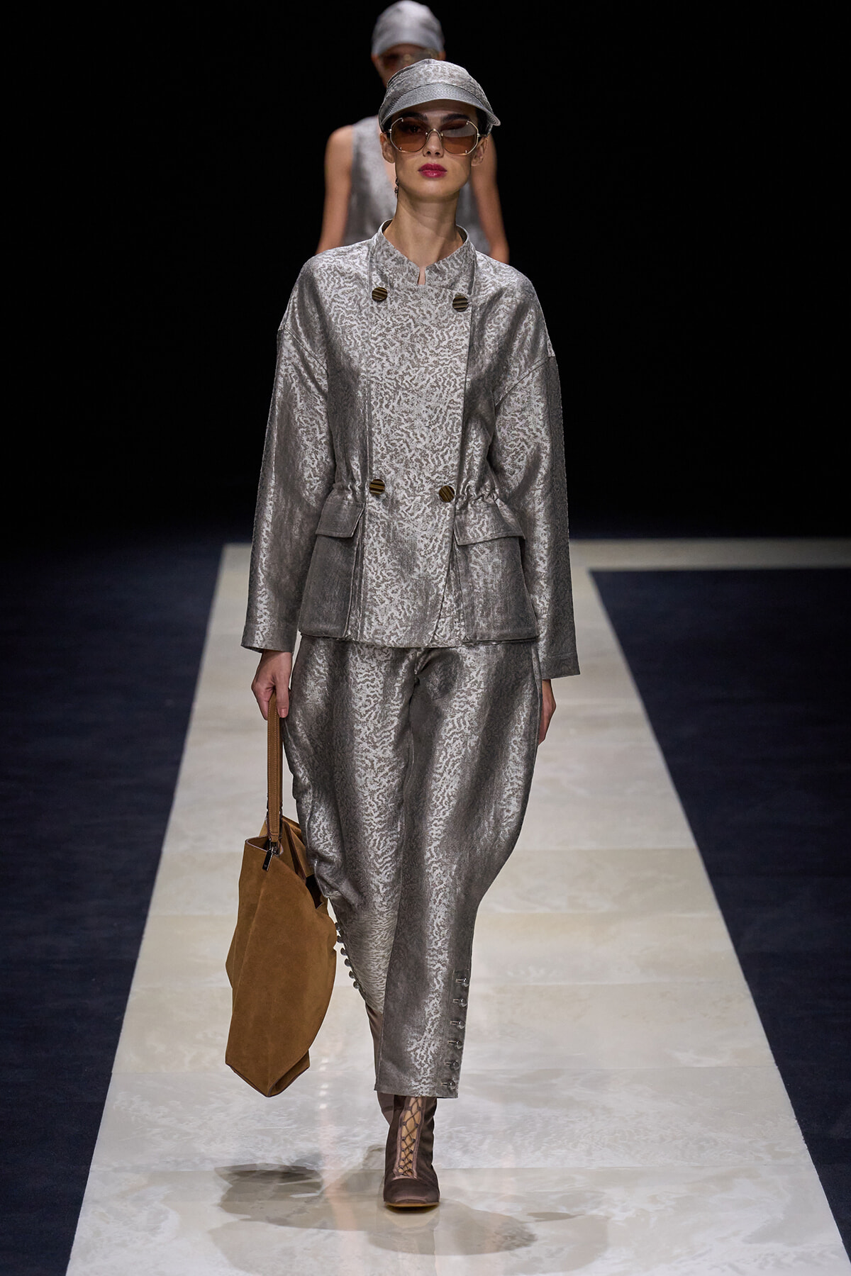 Model on a runway wearing a metallic silver textured suit with cap, sunglasses, and a brown handbag of tan suede kat.