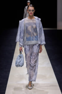 Model on a runway wearing a light blue satin-like jacket and matching wide pants, with a blue graphic top and silver sunglasses, carrying a pale blue drawstring bag.