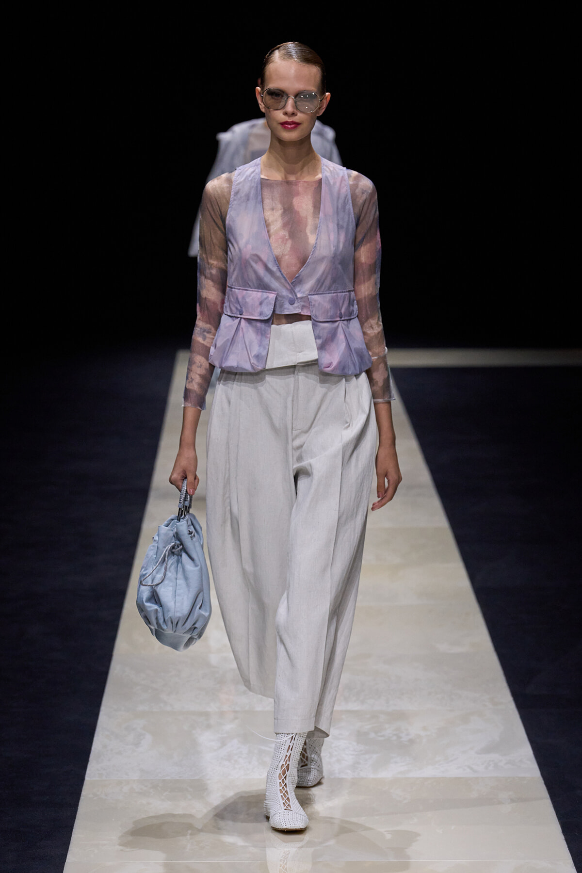 Fashion model on a runway wearing a lavender sheer vest over a pink top, with white wide-leg pants, white lattice boots, and a light blue handbag.