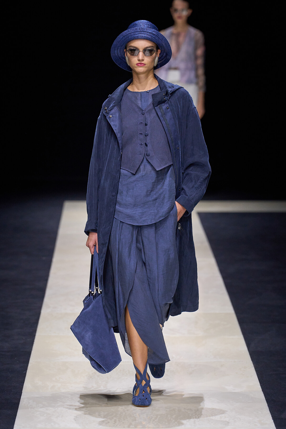 Model walks a runway in an all-blue ensemble: wide-brim hat, trench coat, layered vest, and flowing skirt.