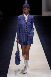 Model walking a runway in a cobalt-blue plaid suit with matching shorts, white scarf, knit cap, sunglasses, and a blue handbag.