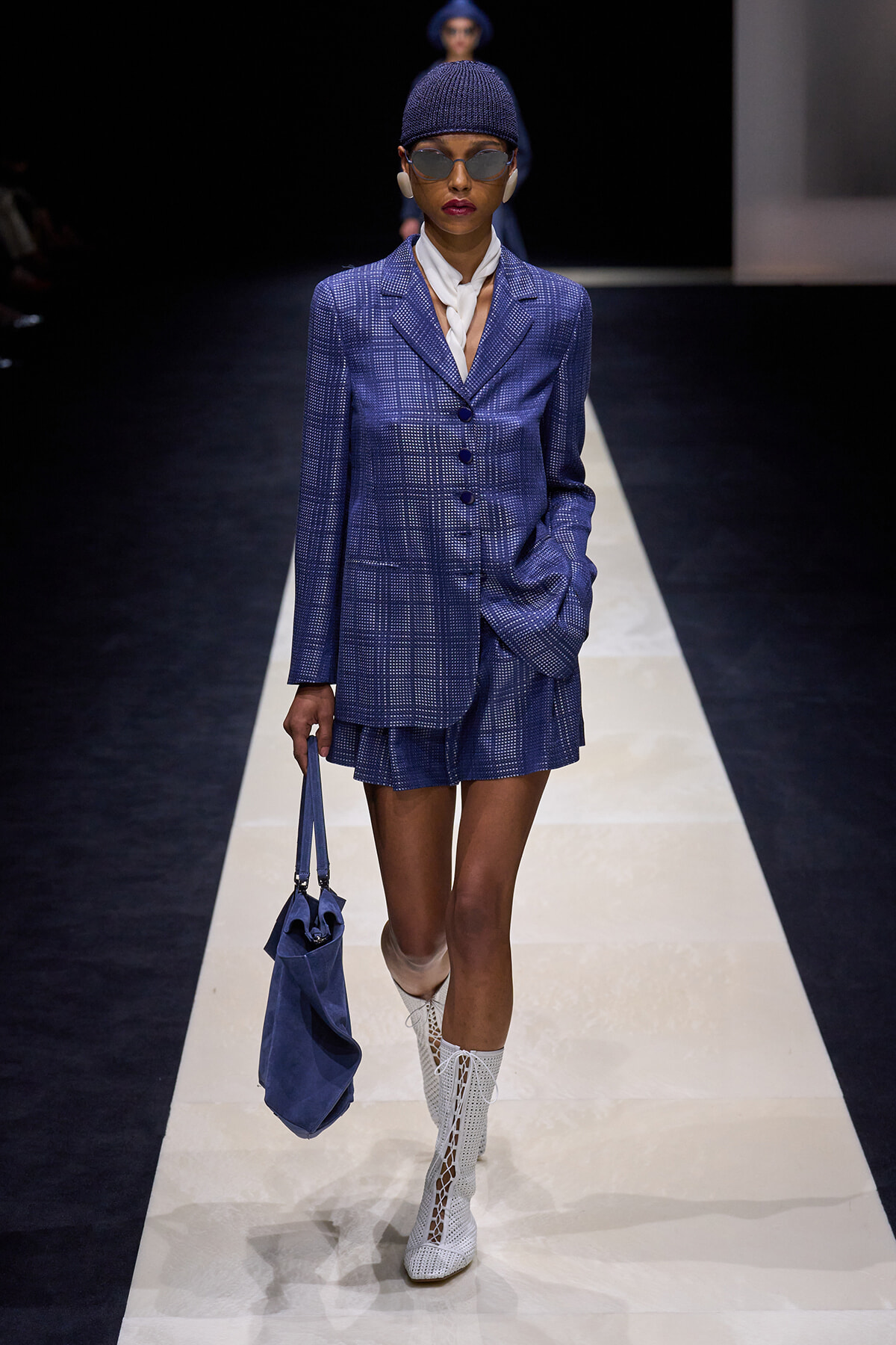 Model walking a runway in a cobalt-blue plaid suit with matching shorts, white scarf, knit cap, sunglasses, and a blue handbag.