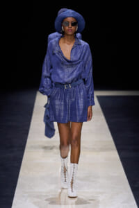 Model walking a fashion runway in a blue metallic shirt dress with a matching belt, wide-brim hat, sunglasses, and white lace-up boots.