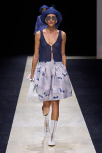 Model on a runway wearing a navy crochet sleeveless top, light blue floral skirt, white fringe bag, and white knee-high boots with a blue hat and sunglasses.
