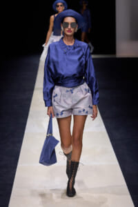 Model walking the runway in a cobalt blue satin blouse with matching hat, light patterned shorts, and knee-high lace-up boots, carrying a blue handbag and wearing sunglasses.
