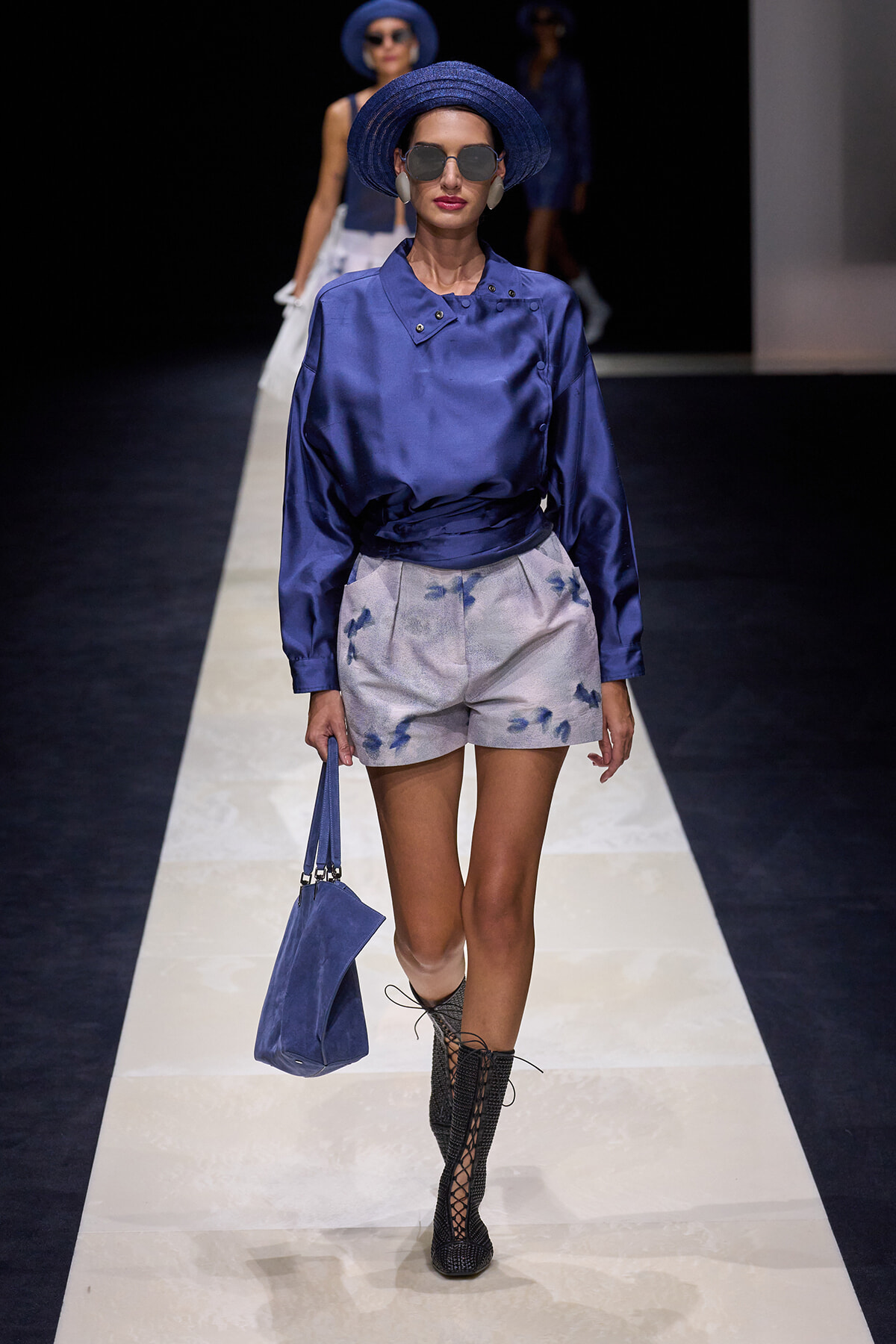 Model walking the runway in a cobalt blue satin blouse with matching hat, light patterned shorts, and knee-high lace-up boots, carrying a blue handbag and wearing sunglasses.