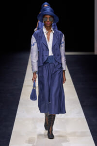 Model on runway in cobalt blue two-piece suit with white blouse, matching hat, and patterned boots.