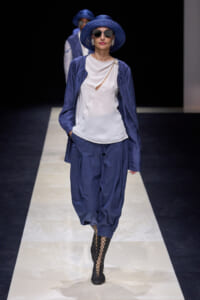 Model on a runway wearing a cobalt blue hat and sunglasses, a white top with a small cutout, a blue jacket, matching blue trousers, and black lattice boots.