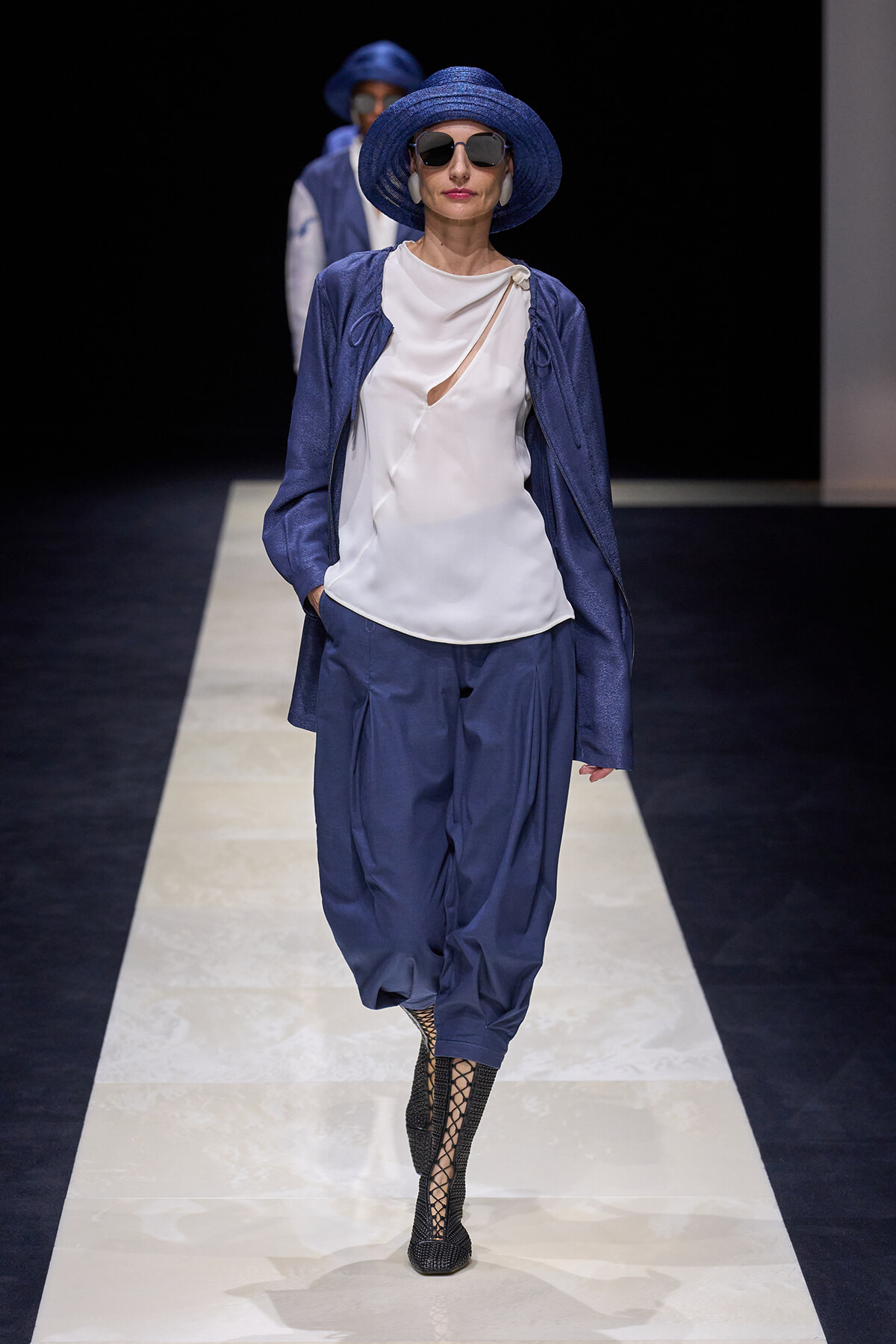 Model on a runway wearing a cobalt blue hat and sunglasses, a white top with a small cutout, a blue jacket, matching blue trousers, and black lattice boots.