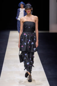 Model walking the runway in a strapless black dress with a floral pattern, cinched waist with a thick woven belt, paired with matching patterned pants and black cutout heels; she carries a small black handbag and wears a black cap and statement earrings.