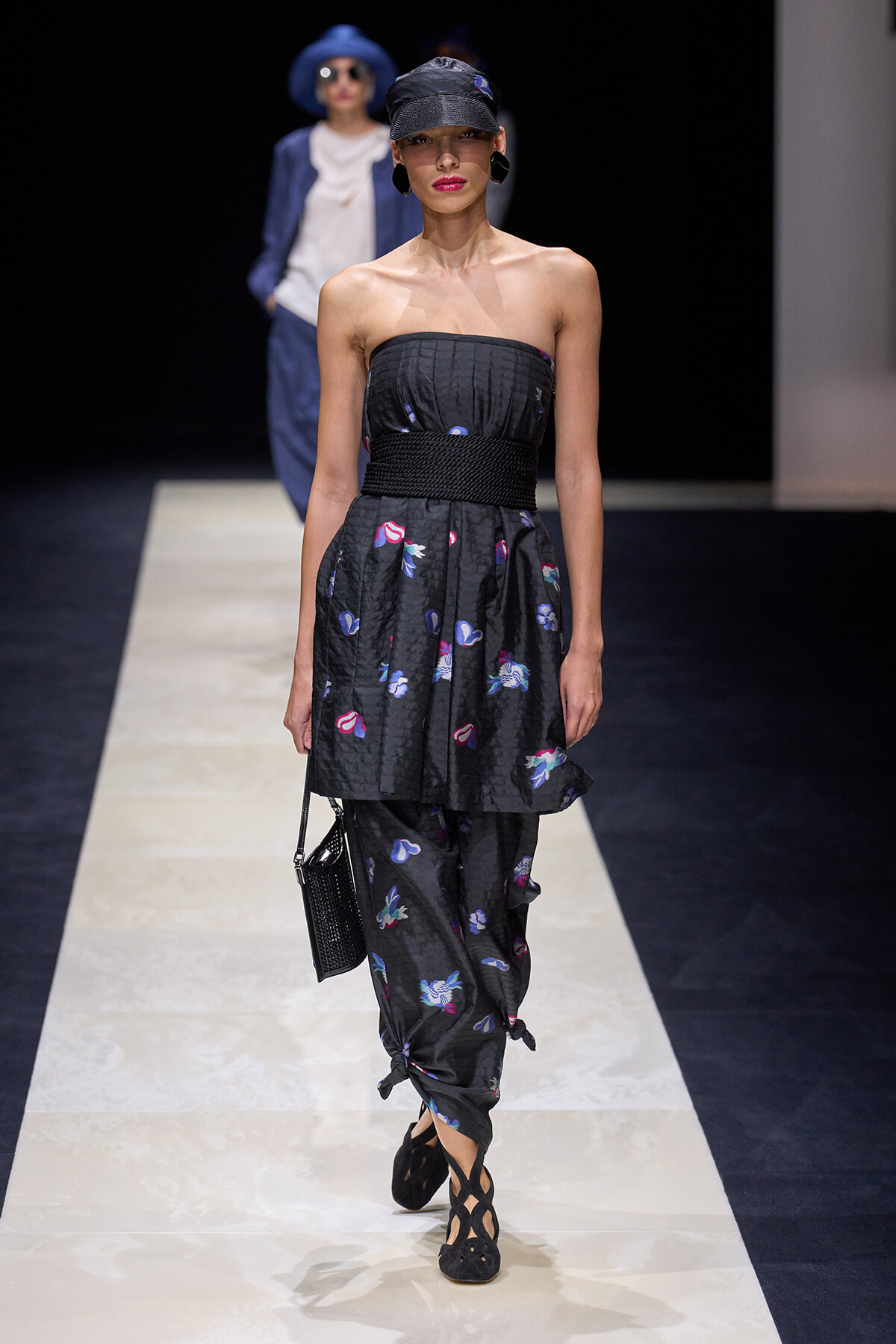 Model walking the runway in a strapless black dress with a floral pattern, cinched waist with a thick woven belt, paired with matching patterned pants and black cutout heels; she carries a small black handbag and wears a black cap and statement earrings.