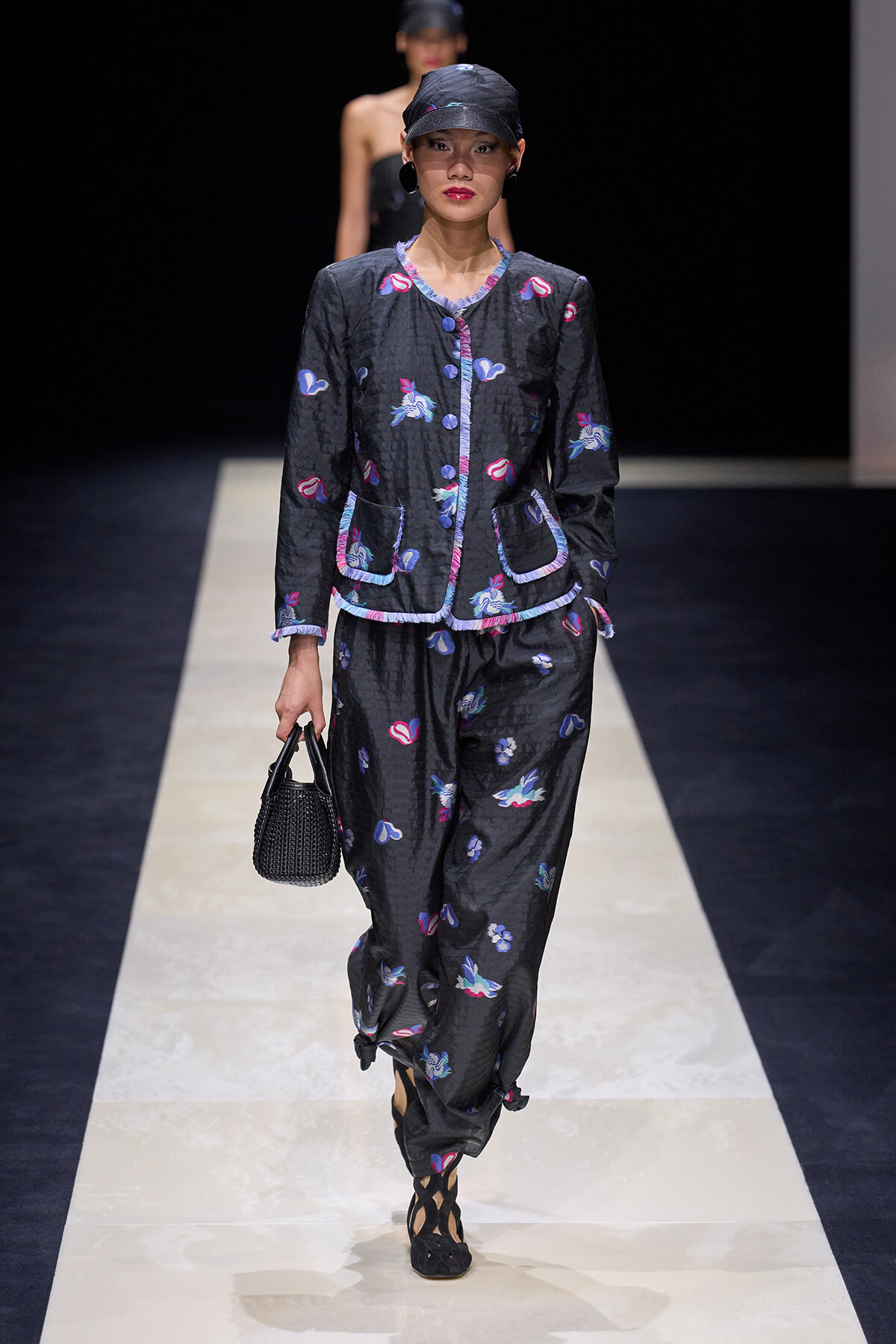 Model walking the runway in a dark quilted two-piece suit with colorful embroidered motifs, matching cap, and a small black woven handbag.
