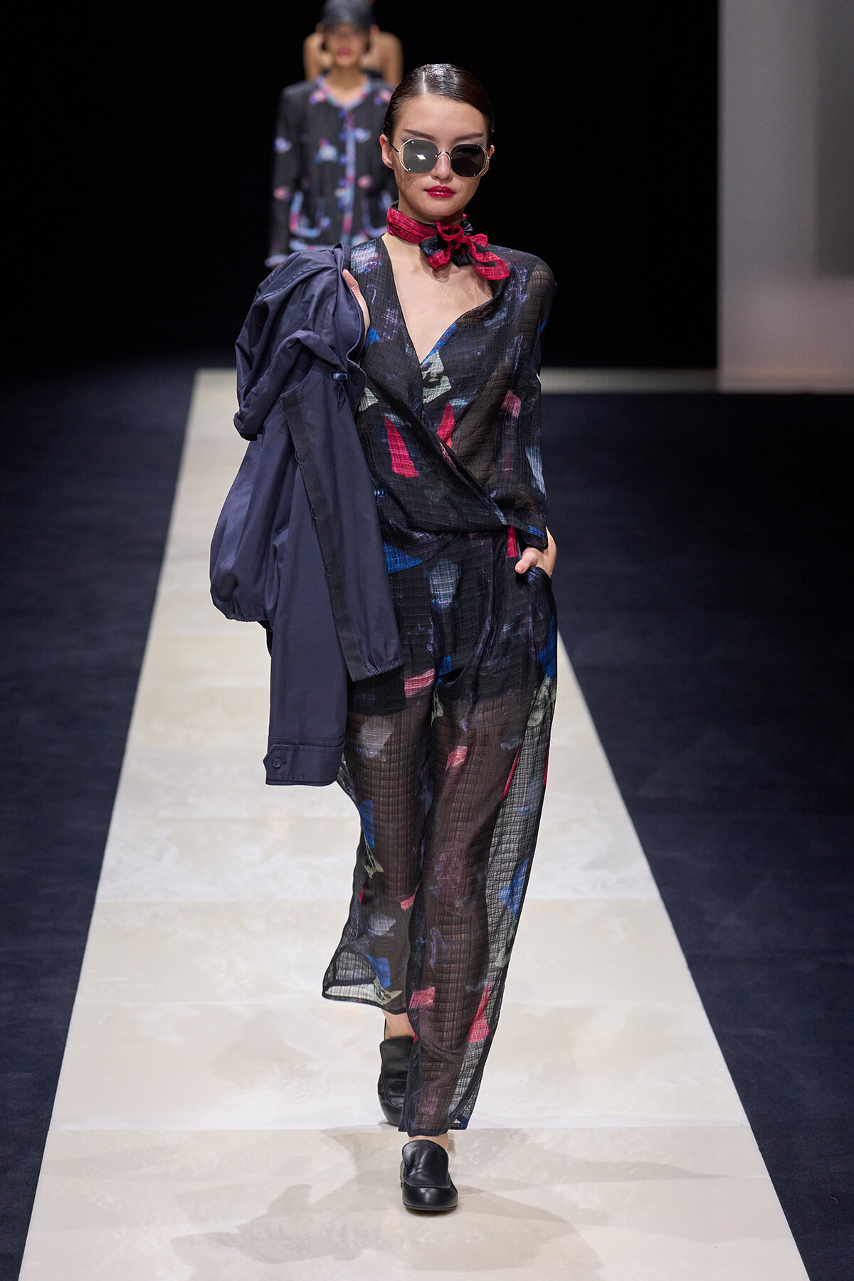 Model walks the runway in a sheer black printed pant suit with blue and red accents, a red neck scarf, sunglasses, and a navy jacket draped over her shoulder.