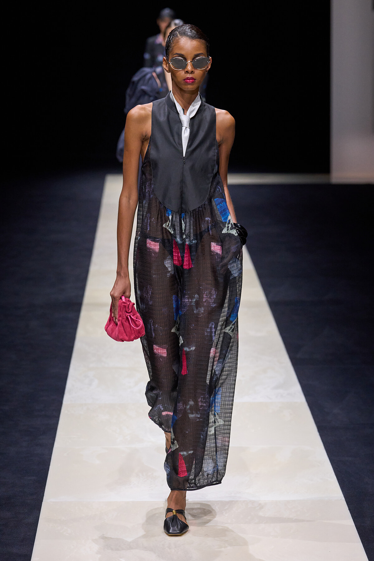 Model walking a runway in a sheer, printed jumpsuit over a black vest with a white necktie, pink clutch in hand, and round sunglasses worn