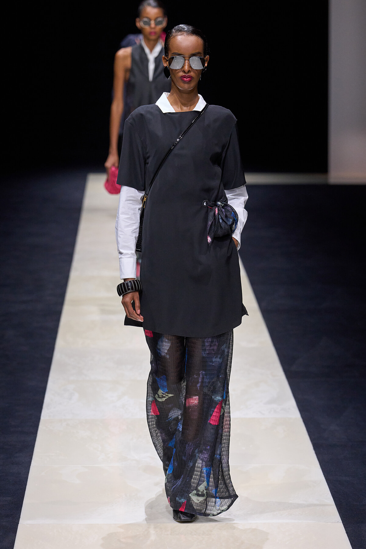 Model walks the runway in a black tunic over a white shirt, paired with sheer patterned pants and a crossbody bag, wearing sunglasses.