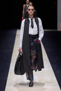 Model walking a fashion runway in a white button-down shirt, black sleeveless vest, tie, and sheer patterned trousers, carrying a black textured tote bag.