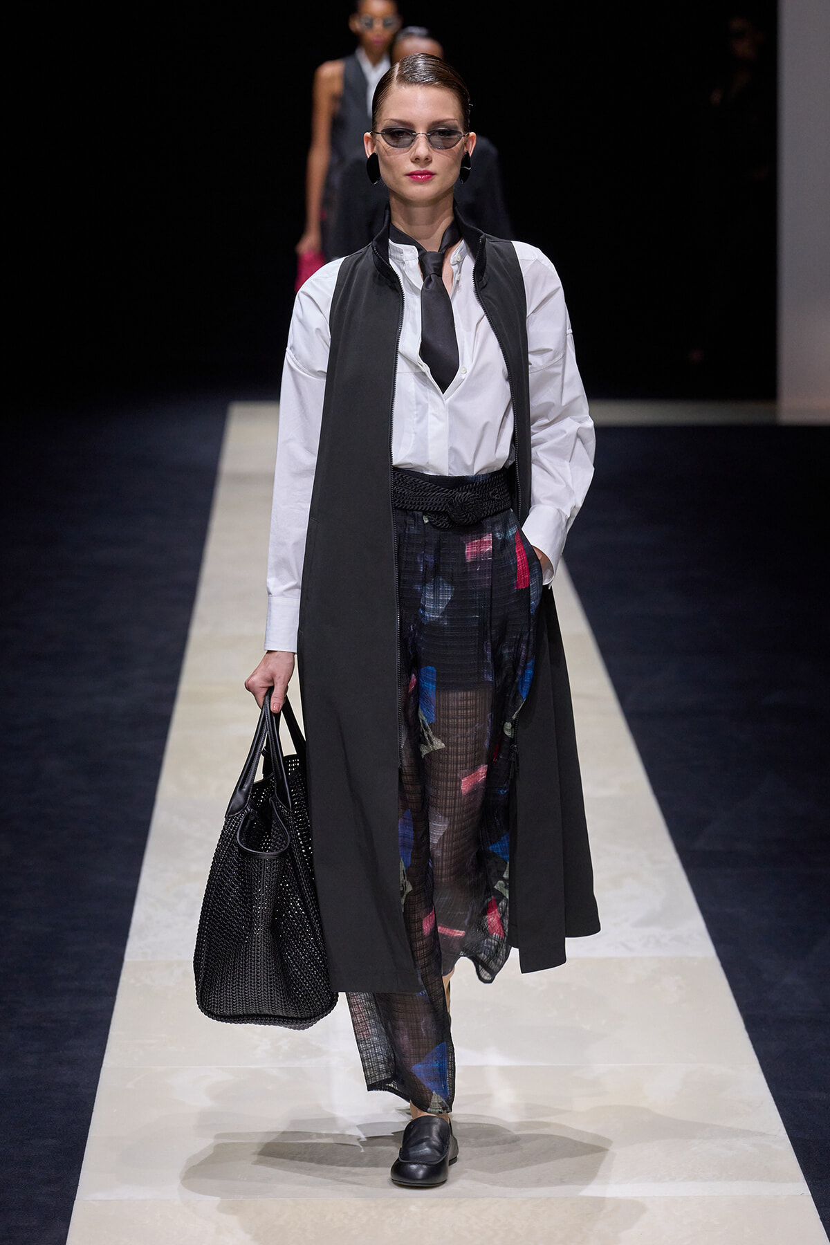 Model walking a fashion runway in a white button-down shirt, black sleeveless vest, tie, and sheer patterned trousers, carrying a black textured tote bag.