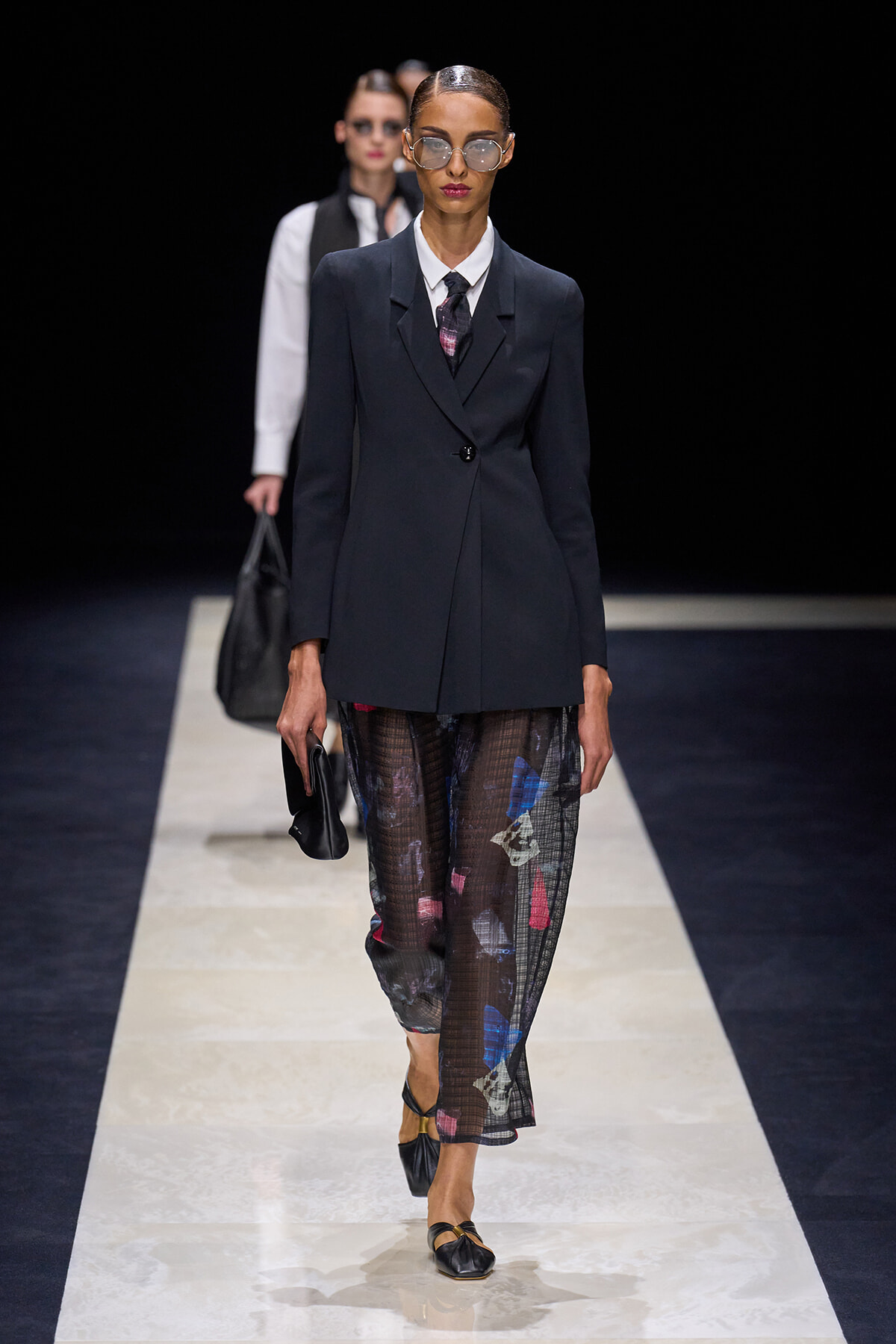 Model walks a runway in a dark tailored blazer, white shirt with tie, and sheer printed trousers with black flats and sunglasses variant sunglasses and clutch in hand.